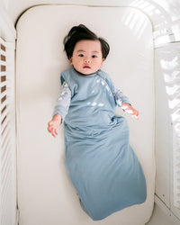 A baby lying on their back inside a white crib, wearing a light blue swaddle blanket over a long-sleeve light gray shirt. The crib has a white mattress and white slatted sides, with soft natural light casting shadows on the right side.