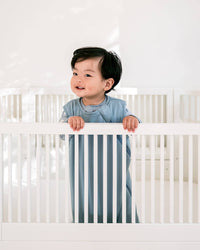 A baby wearing a light blue long-sleeve top stands behind a white crib in a bright, minimalist room.