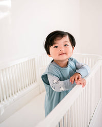 A baby wearing a light blue long-sleeve sleep sack with patterned sleeves, leaning on a white crib in a bright, minimalist nursery.