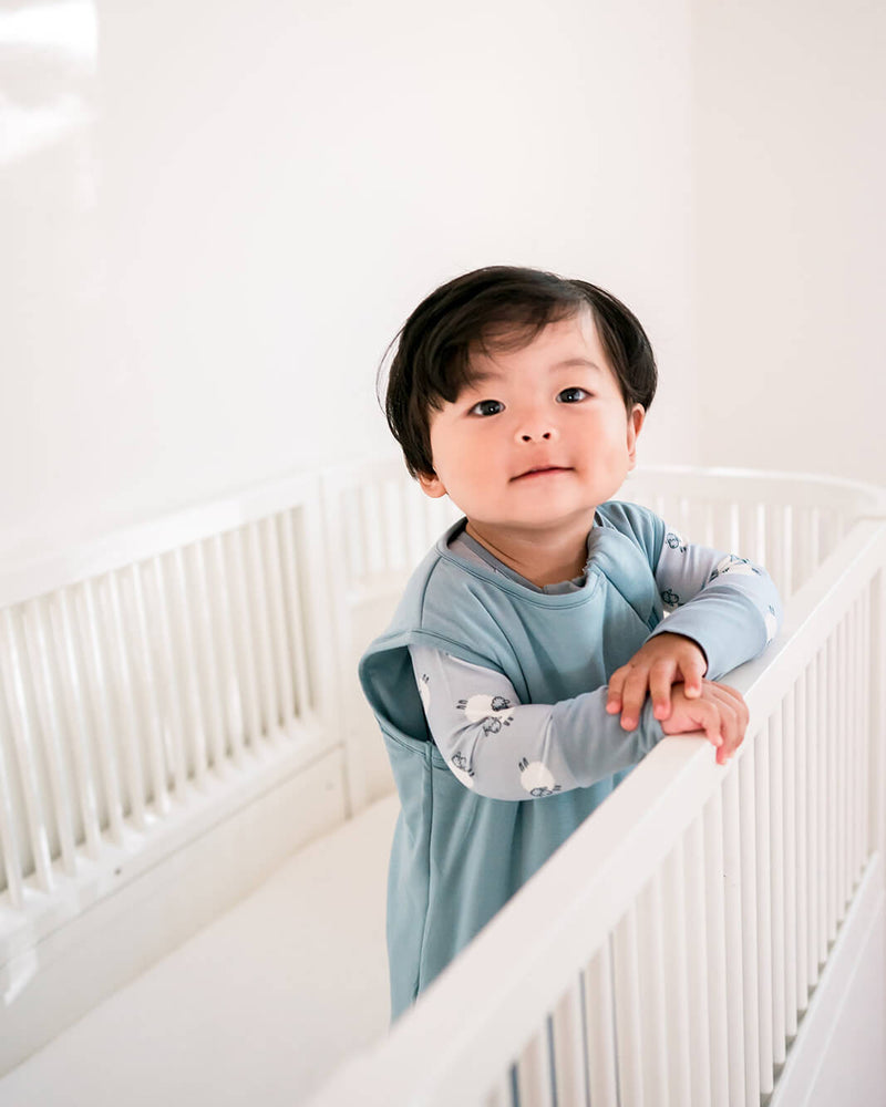A baby wearing a light blue long-sleeve sleep sack with patterned sleeves, leaning on a white crib in a bright, minimalist nursery.
