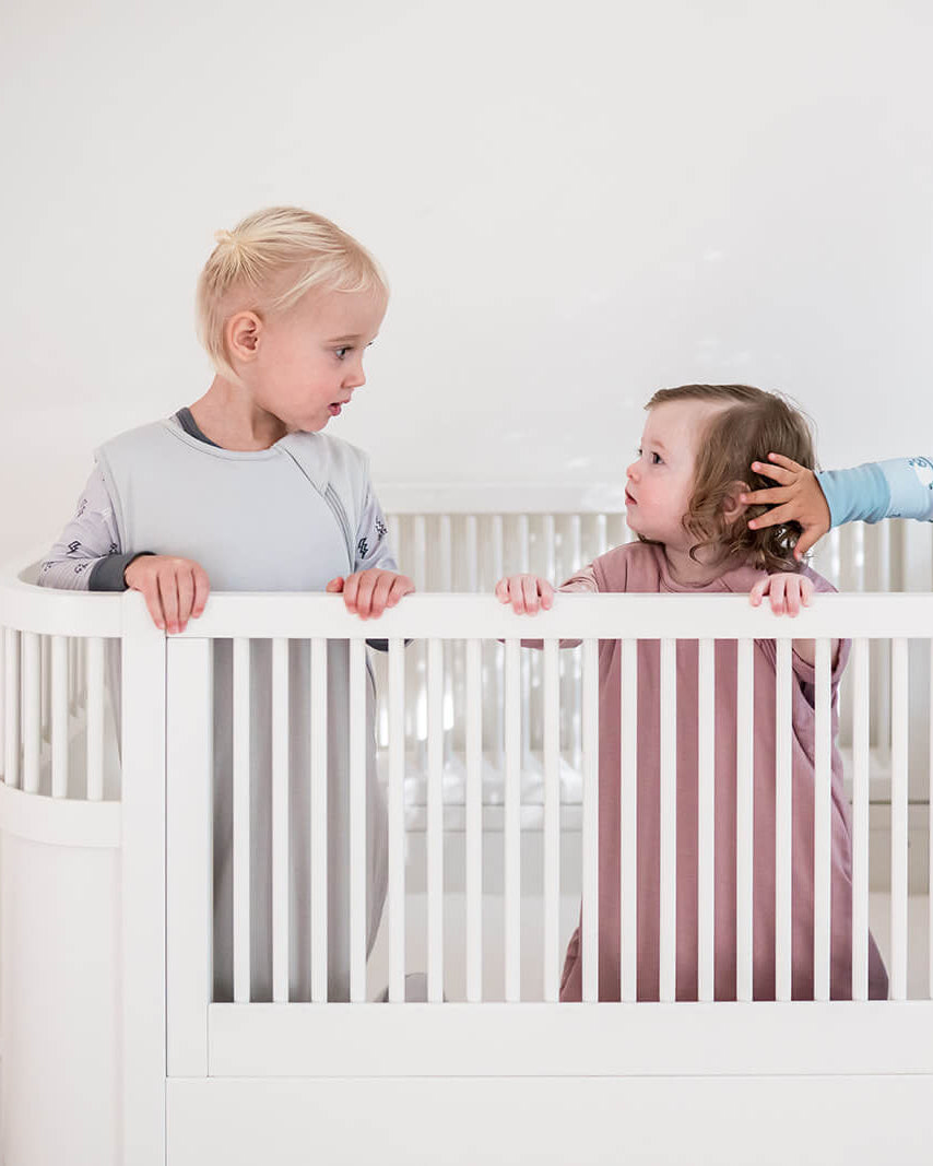 Three young children standing in a white crib or playpen against a plain white wall, wearing light-colored pajamas in a bright, minimalist room.