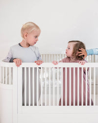 Three young children standing in a white crib or playpen against a plain white wall, wearing light-colored pajamas in a bright, minimalist room.