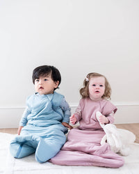 Two toddlers sitting on a white rug in a bright room, wearing matching footed sleepers in light blue and dusty pink. The child on the right holds a white plush toy.