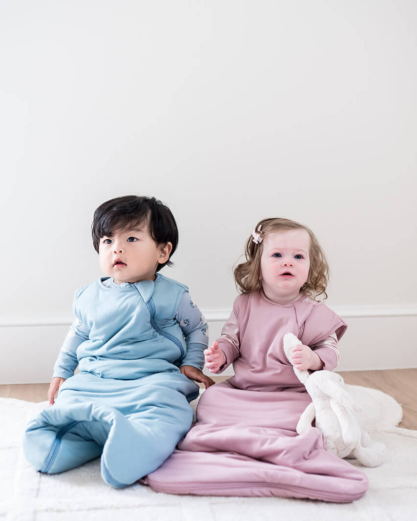 Two toddlers sitting on a white rug in a bright room, wearing matching footed sleepers in light blue and dusty pink. The child on the right holds a white plush toy.