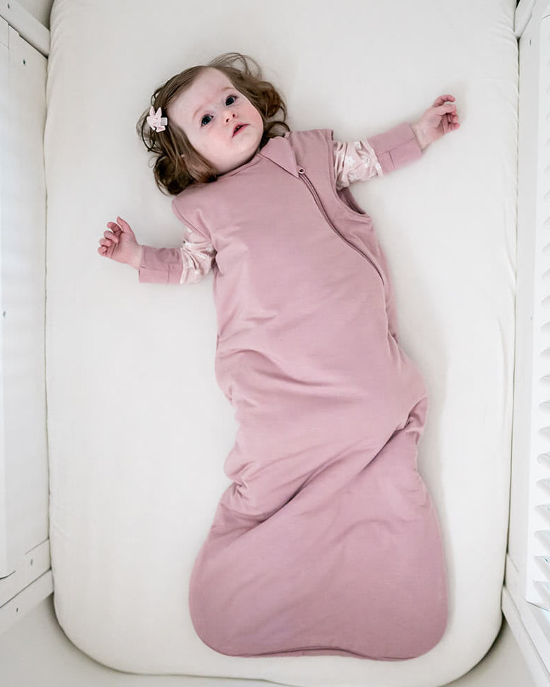 A baby wearing a dusty pink sleep sack lies on their back inside a white wooden crib with a white mattress.