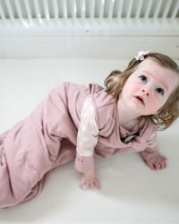 A baby girl wearing a dusty pink long-sleeve sleep sack with white lace details on the sleeves, crawling on a white crib mattress.