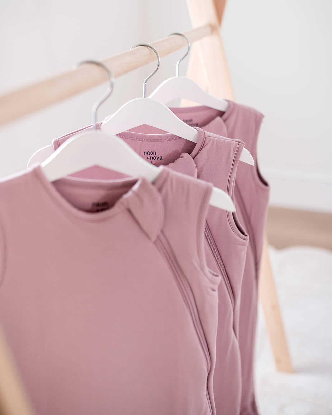 A close-up of several sleeveless dusty pink tops hanging on white hangers from a light wood clothing rack in a bright, softly lit room.