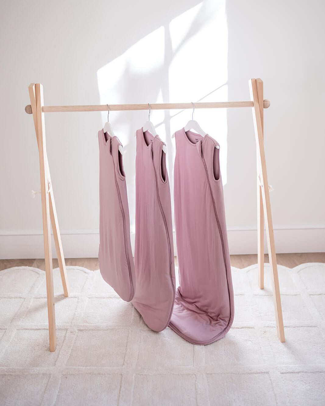 Three dusty pink sleeveless baby blankets hanging on a light wood A-frame rack on a white textured rug against a white wall with natural window light.