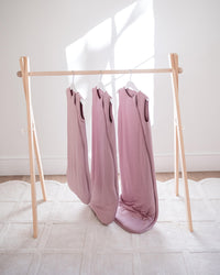 Three dusty pink sleeveless baby blankets hanging on a light wood A-frame rack on a white textured rug against a white wall with natural window light.