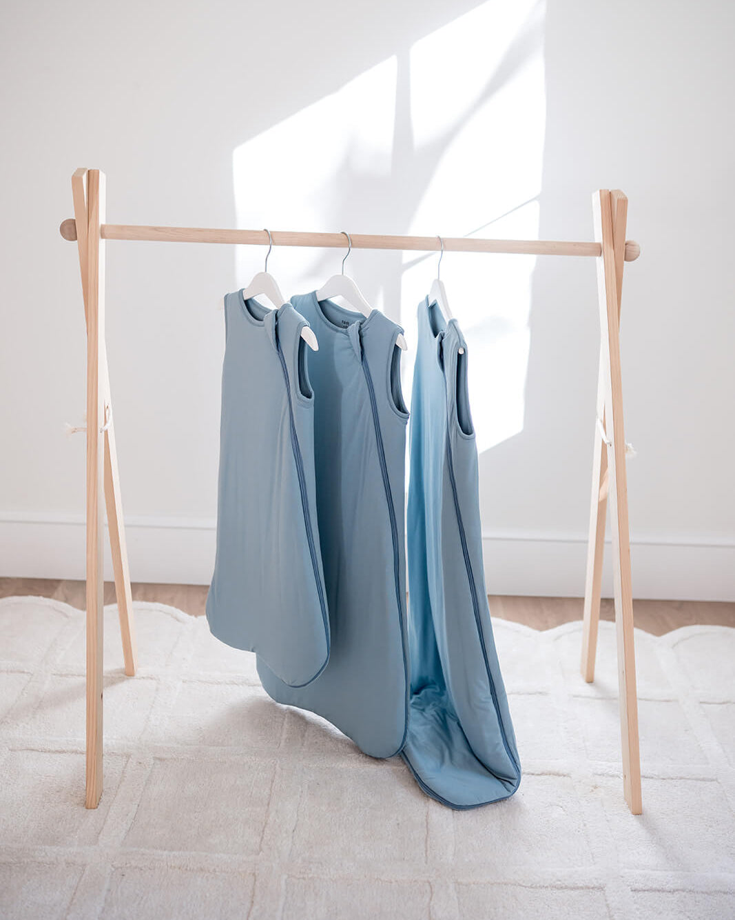 Three light blue sleeveless baby garments hanging on a wooden A-frame clothing rack in a bright, minimalist room with natural sunlight casting shadows on the wall.