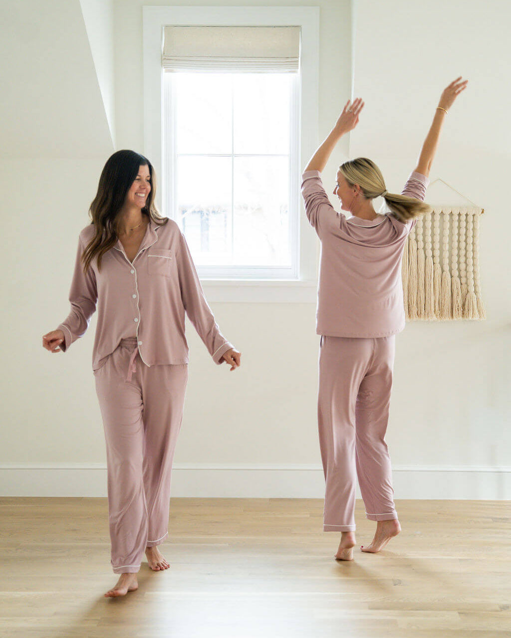 Two women in matching pink pajama sets standing in a room with a window.