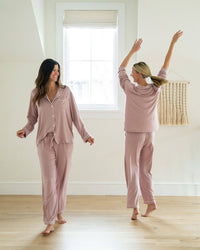 Two women in matching pink pajama sets standing in a room with a window.