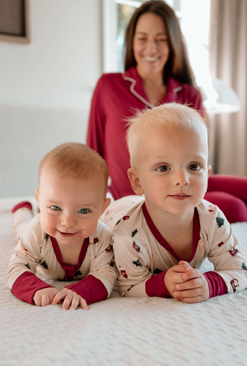 Two toddlers lying on a bed wearing matching cream and burgundy pajamas with a small holiday print, with a smiling woman in matching burgundy pajamas standing in the background.