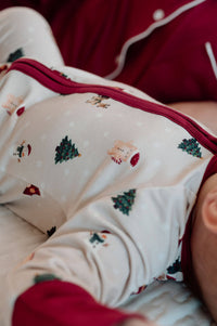 A close-up of a baby wearing a cream-colored Christmas-themed long-sleeve pajama top with red trim and holiday prints, lying next to an adult in a dark red pajama set.