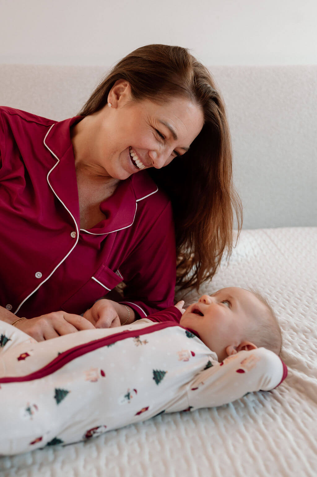 A woman in a red pajama with white piping smiles while lying on a bed with a baby wearing a white Christmas-themed footed sleeper.