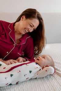 A woman in a red pajama with white piping smiles while lying on a bed with a baby wearing a white Christmas-themed footed sleeper.