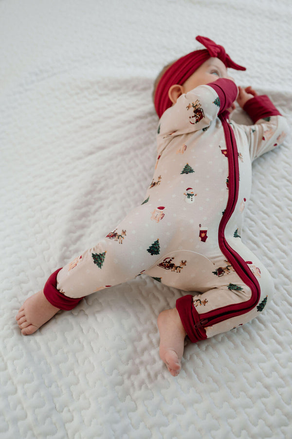 A baby lying on a white textured blanket wearing a festive Christmas print footless sleeper with red trim, a red bow headband, and red mittens.