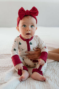 A baby wearing a festive holiday-themed footed sleeper and a matching red bow headband, sitting on a white quilted blanket while being held.